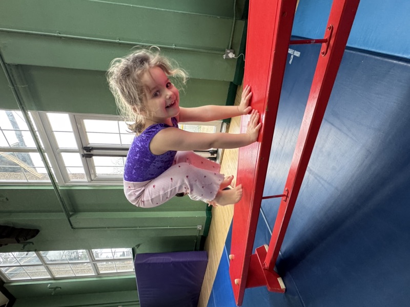 A young girl smiling as she climbs the wall bars at TJ's Gymnastics Club